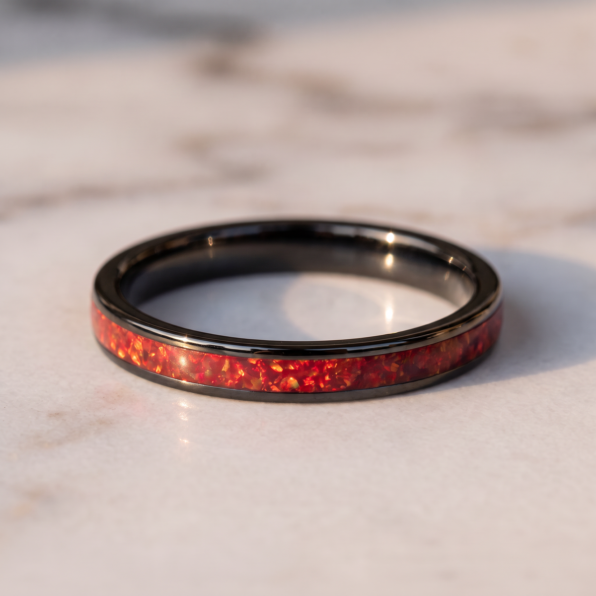 The Cinder black tungsten ring with a red opal inlay lying flat on a marble surface