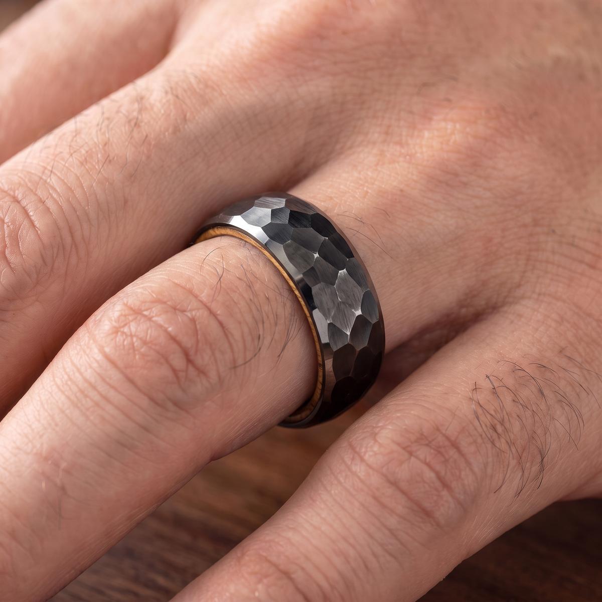 Close up of a man's hand wearing The Cask black hammered tungsten ring with a whiskey barrel wood interior
