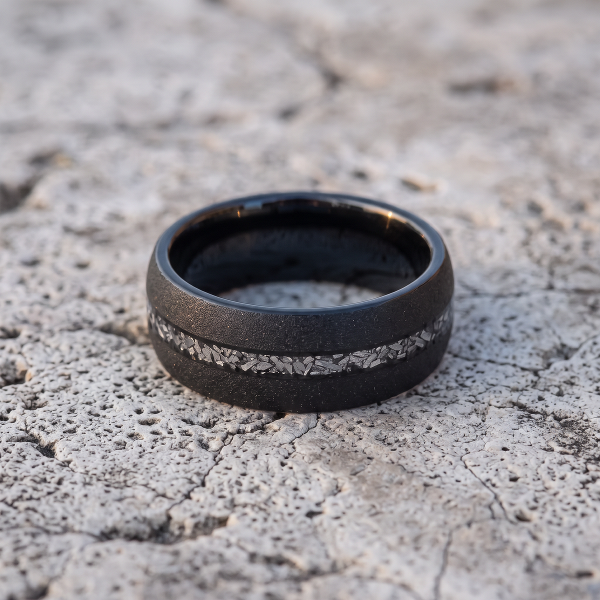The Stardust black sandblasted tungsten ring with meteorite inlay lying flat on light grey stone