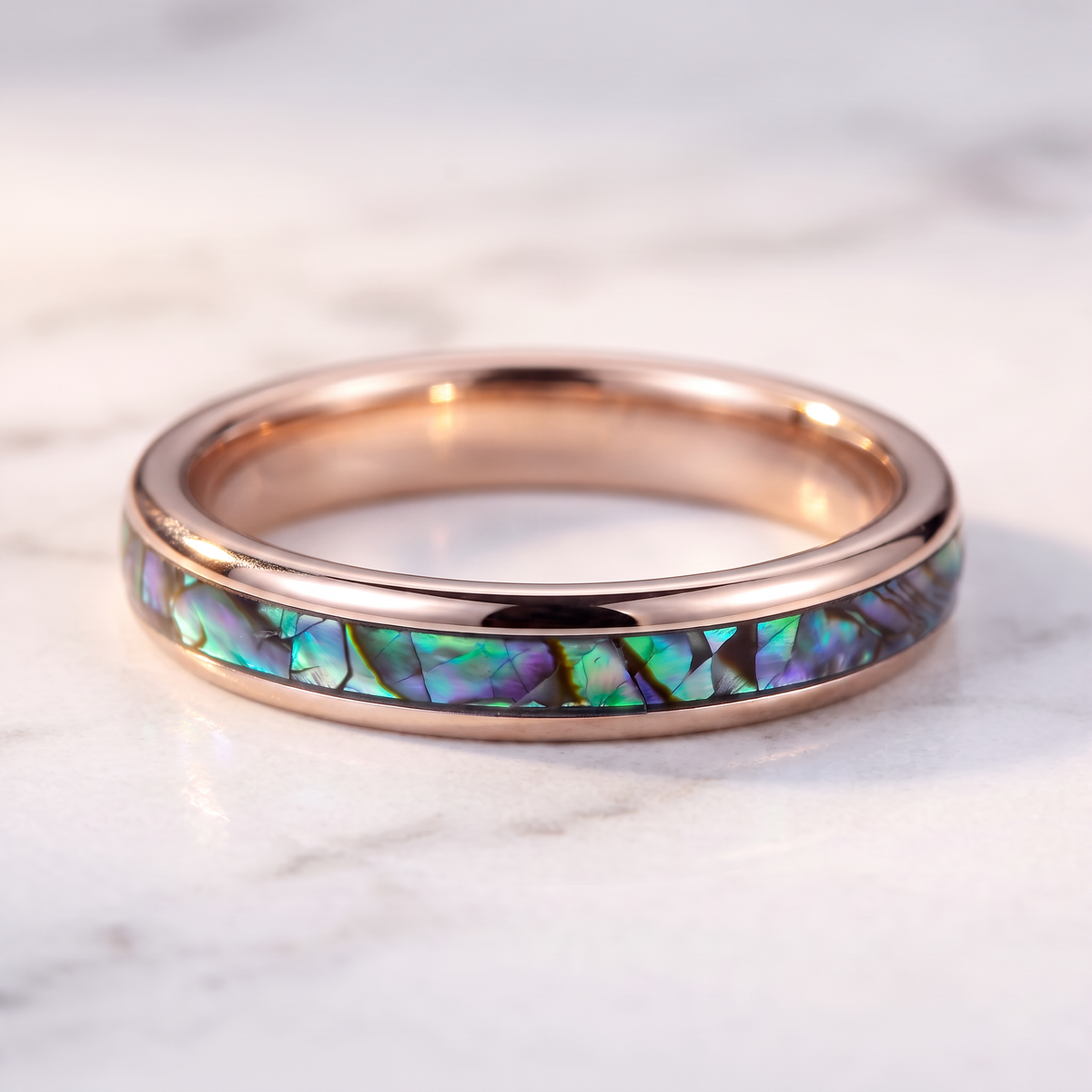 The Iris rose color tungsten ring with an abalone shell inlay lying flat on a marble surface