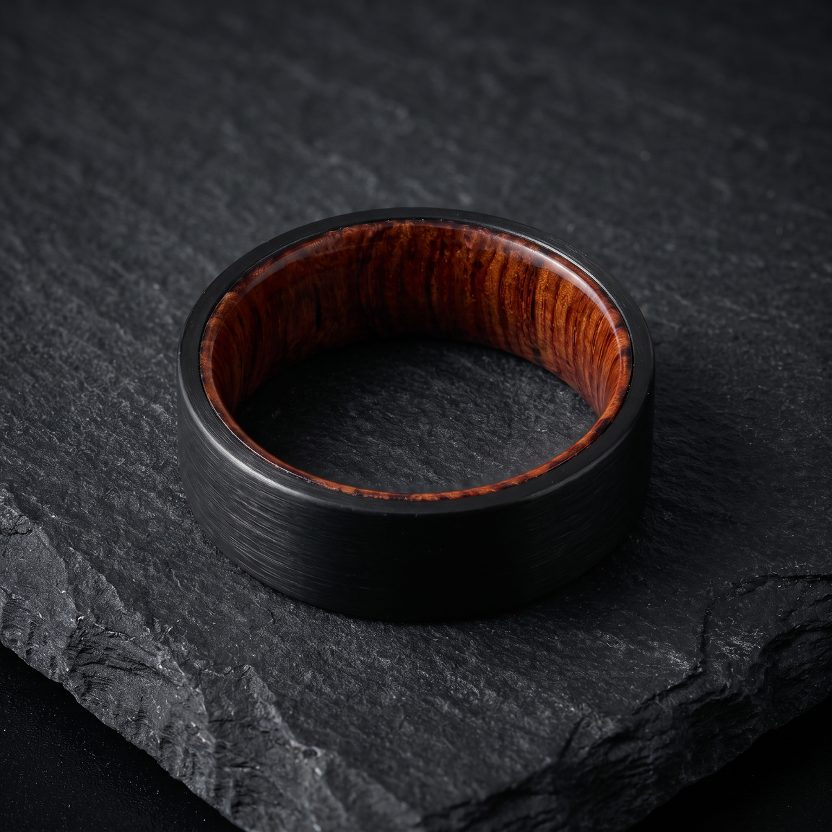 The Ember tungsten ring with a koa wood interior lying flat on a dark slate surface