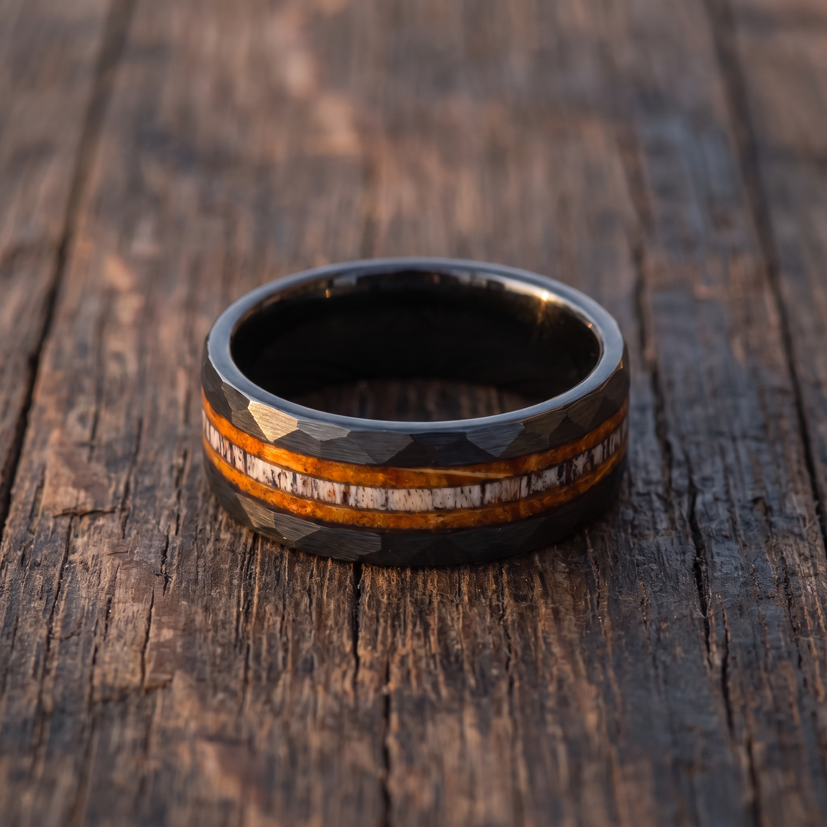 The Cooper tungsten ring with whiskey barrel wood and deer antler inlay lying flat on a weathered dark oak surface