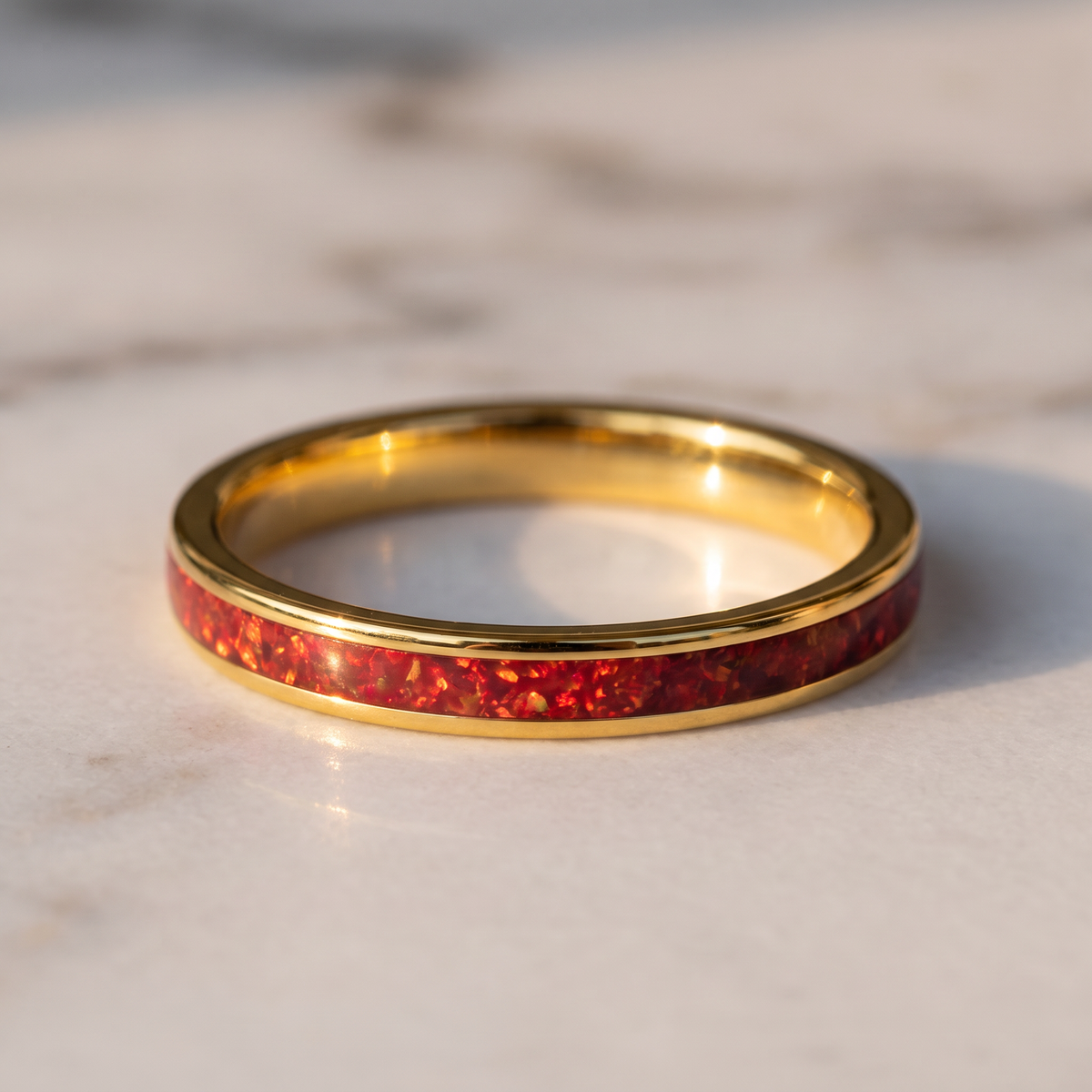 The Cinder gold color tungsten ring with a red opal inlay lying flat on a marble surface