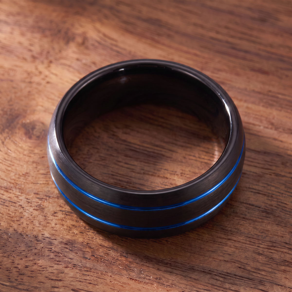 The Riptide black tungsten ring with a double blue line inlay lying flat on dark walnut wood