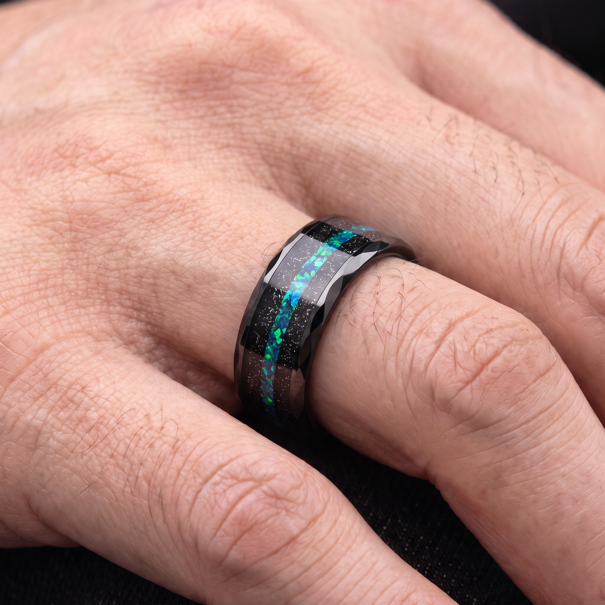 Close up of a man's hand wearing The Reef black tungsten ring with black sand and green blue opal inlay